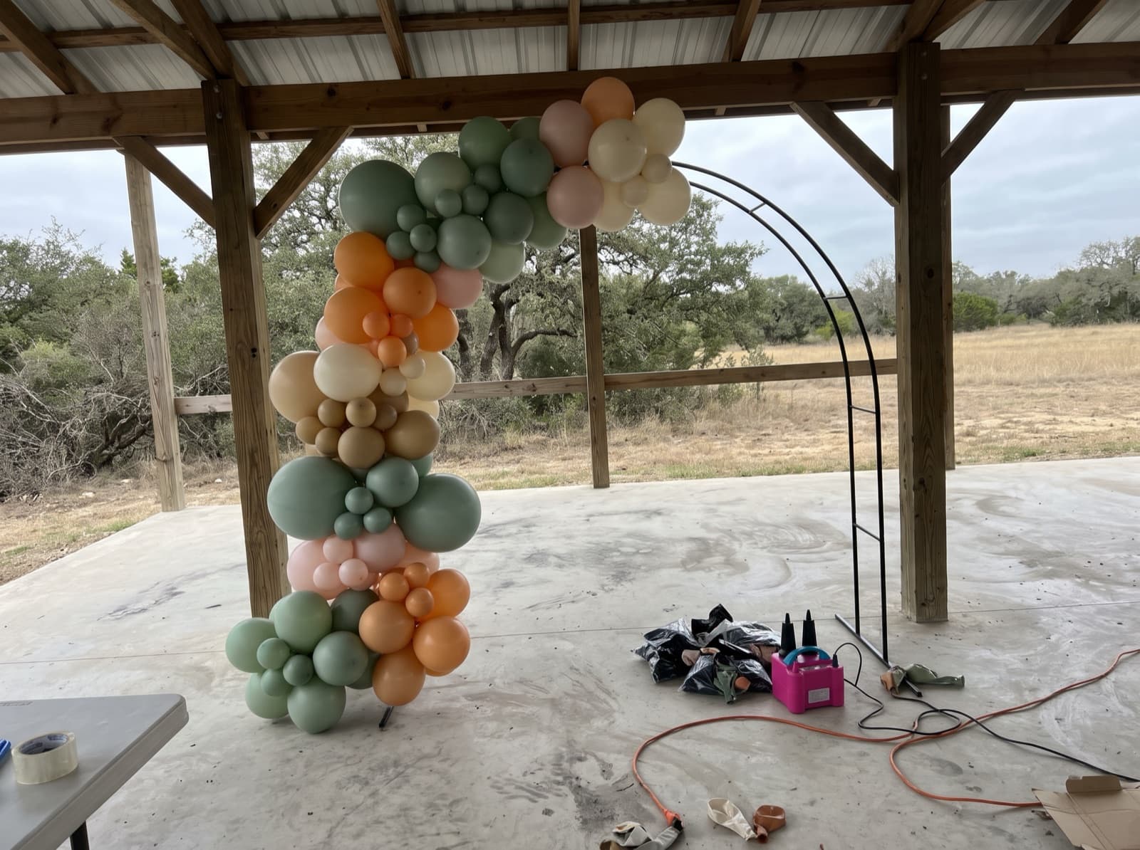 Organic balloon garland half-built on an arch frame inside an open-air ranch pavilion